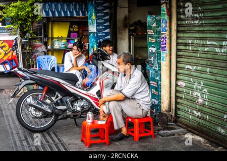 Three Vietnamese people enjoying their morning coffee in front of a garage door sitting on short stools. Stock Photo