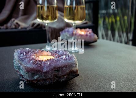 Selective focus on amethyst crystal geode candle holder with tea light candle burning inside, two white wine glasses on background in the evening. Stock Photo
