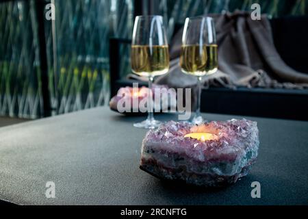 Selective focus on amethyst crystal geode candle holder with tea light candle burning inside, two white wine glasses on background in the evening. Stock Photo