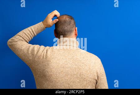 Back view of thoughtful man scratching his head over blue background. He wonders why he doesn't have luck in life and if he will always be like this Stock Photo