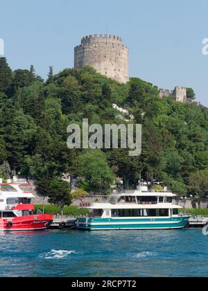 Rumeli Fortress surrounded by trees on a hill on the banks of the Bosphorus Sea with passenger ferries docked, Istanbul, Turkey Stock Photo