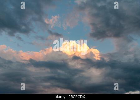 Opening in dark thunderclouds with blue sky and sun coloring white cumulus clouds orange during sunset Stock Photo