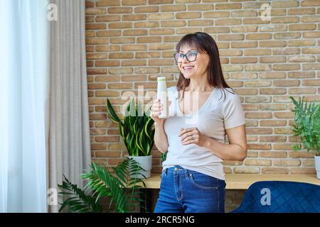 Middle aged woman drinking fermented milk product from bottle, near window Stock Photo