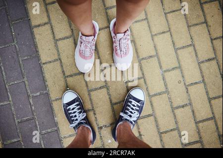 View from above of male and female legs in pink and blue sneakers, standing on the pavement. Selective focus. Close-up. Copy advertising space Stock Photo