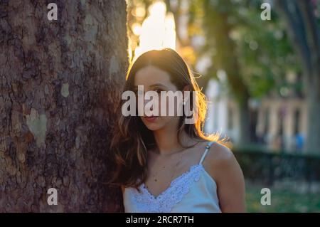 Horizontal close up shot of a pretty young woman standing beside a tree looking at the camera Stock Photo
