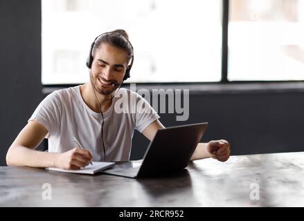 Guy Taking Notes At Laptop Posing With Headset In Office Stock Photo