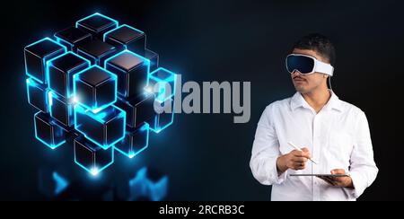 Young man wearing innovative vision pro virtual reality glasses and using digital device, blue glowing holographic cubes Stock Photo
