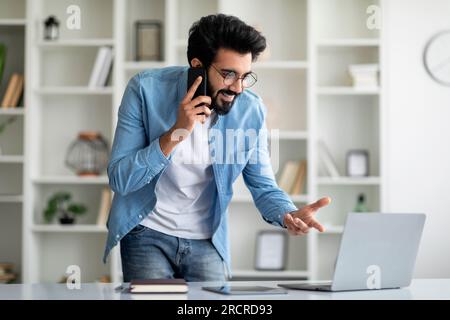 Smiling Indian Freelancer Guy Wotking With Cellphone And Laptop At Home Office Stock Photo
