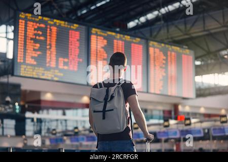 Traveling by airplane. Man walking with backpack and suitcase walking through airport terminal and looking at departure information. Stock Photo