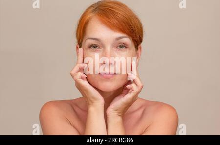 Portrait of caucasian middle aged woman of 40s with reddish hair looking at camera holding fingers on cheek , showing soft skin Stock Photo