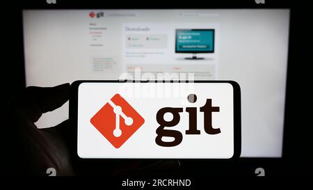 Person holding mobile phone with logo of distributed version control system Git on screen in front of web page. Focus on phone display. Stock Photo