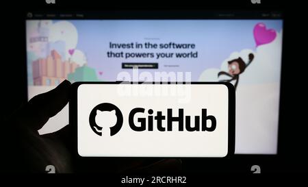 Person holding smartphone with logo of US software development company GitHub Inc. on screen in front of website. Focus on phone display. Stock Photo