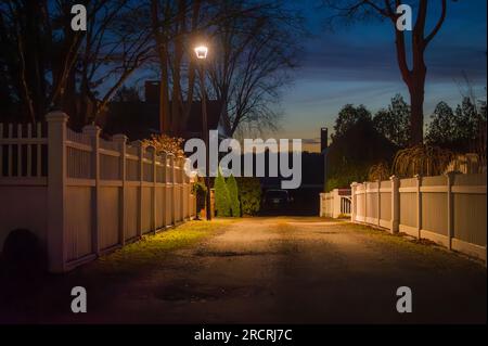 Experience the magic of twilight as a streetlamp guides you past picturesque white fences in this New England village. Every detail whispers a story. Stock Photo