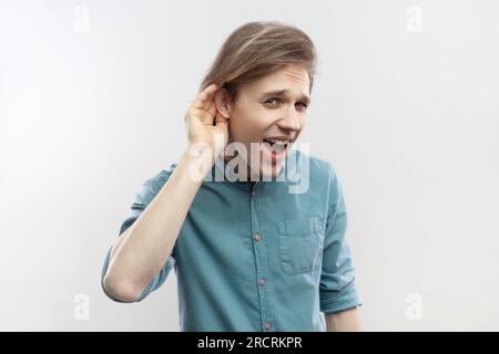 What I dont hear you. Portrait of man standing with hand near ear and listening to whisper, difficult to understand quiet talk, wearing blue shirt. Indoor studio shot isolated on gray background. Stock Photo