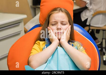 Schoolgirl experiencing severe toothache while sitting in dental chair before appointment with dentist. Girl on dental appointment. Regular check up t Stock Photo