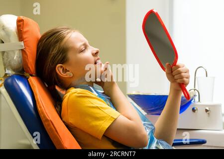 Schoolgirl looking in mirror and experiencing severe toothache while sitting in dental chair before appointment with dentist. Girl on dental appointme Stock Photo