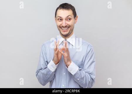 Portrait of cunning sly young adult man standing looking away with toothy smile, having devil plans, planning prank, wearing light blue shirt. Indoor studio shot isolated on gray background. Stock Photo