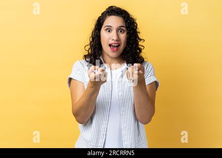 Portrait of amazed excited beautiful woman with dark wavy hair pointing directly at you, choosing something, has shocked expression. Indoor studio shot isolated on yellow background. Stock Photo