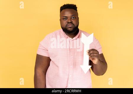 Portrait of sad upset depressed bearded man wearing pink shirt showing white arrow pointing down, expressing sorrow, downgrade concept. Indoor studio shot isolated on yellow background. Stock Photo