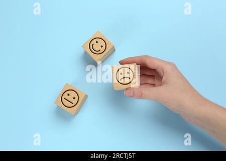 Complaint. Woman choosing wooden cube with sad emoticon instead of other ones with drawn happy and neutral faces on light blue background, top view Stock Photo