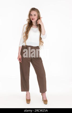 Young blonde woman showing thumbs up and playfully sticks out tongue, looking at camera. Caucasian female dressed in white blouse, brown trousers Stock Photo
