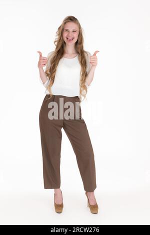 Happiness young woman showing thumbs up with both hands, playfully sticks out tongue. Caucasian blonde standing full length on white background Stock Photo