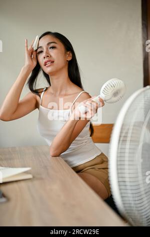A portrait of a beautiful Asian woman feeling overheated, suffering from a heat attack, wiping sweat on her face, and cooling herself down with an ele Stock Photo