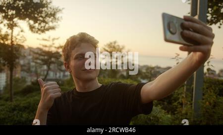 Capturing Moments: Handsome Blond Young Man Taking Selfies with His Smartphone, Striking Various Poses Outdoors Stock Photo