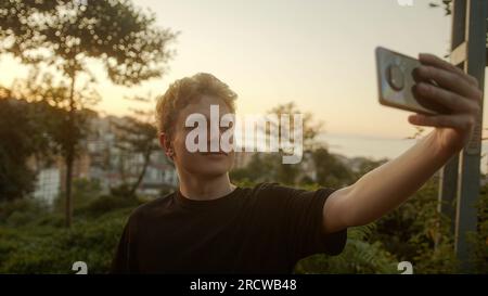 Blond Young Man Taking Selfies with His Smartphone, Striking Various Poses Outdoors Stock Photo