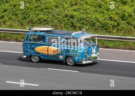 Warren's Vintage VW. 1973 70s seventies Blue Custom VW Volkswagen Motor Caravan LCV Petrol 1584 cc. A curious wedding hire car with hippie decorations, white ribbons & surfboard; travelling at speed on the M6 motorway in Greater Manchester, UK Stock Photo