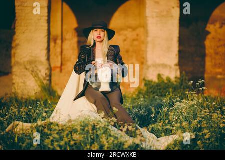 Woman blonde in hat sitting on chair in nature on background of old building with arches. Lady is holding an aquarium and a mannequin head in her hand Stock Photo