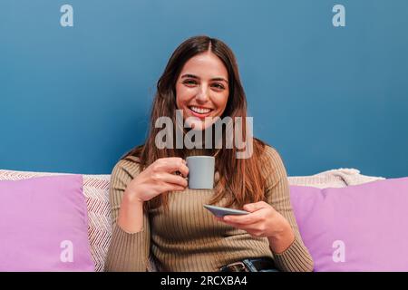 Portrait of caucasian young happy woman posing holding a coffee cup on her hand, smiling and enjoying the moment sitting on the sofa at her cosy apartment . Joyful female drinking a tea at home. High quality photo Stock Photo