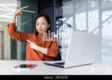 Portrait of a young successful Asian female programmer, developer and IT expert. Sitting at a table and working on a laptop, pointing at the camera with a finger on his hand, a gesture of strength. Stock Photo