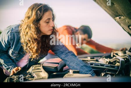 A young couple had a car breakdown on the road. They stand very upset, open the hood and try to fix the broken engine Stock Photo