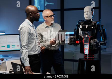 Two multiethnic engineers examining program of robot using digital tablet while standing in office Stock Photo