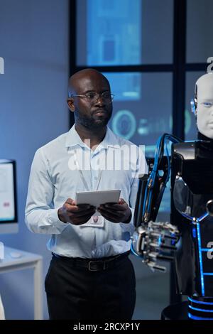 Vertical image of African American developer using tablet pc to control the robot in the lab Stock Photo