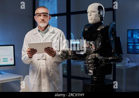 Portrait of mature engineer in white coat looking at camera while working with programming of robot using digital tablet Stock Photo
