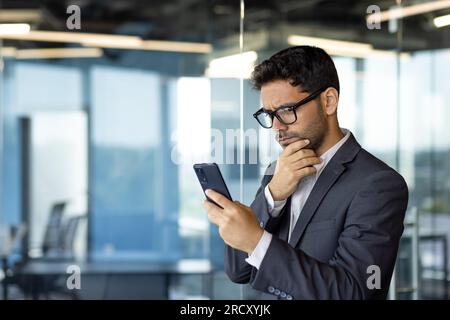 Thinking businessman inside office with phone, Arab in glasses works at workplace, uses application on smartphone online reads and browses internet pages thoughtfully. Stock Photo