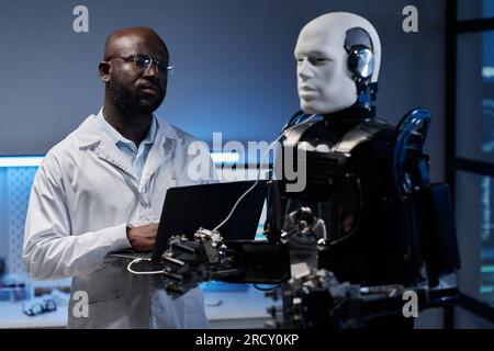 African American engineer in lab coat using laptop to control the robot in office Stock Photo