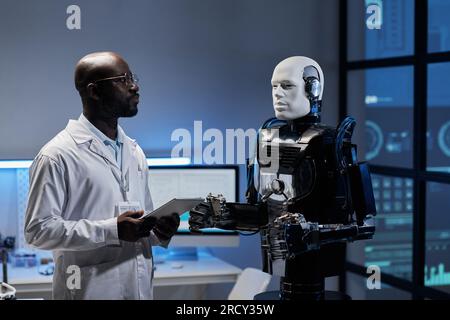 African American engineer in white coat working on tablet pc with robot in office Stock Photo