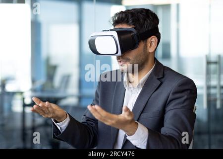 Young arab businessman talking online remotely, using vr glasses, man on remote meeting with colleagues in metaverse, sitting inside office. Stock Photo