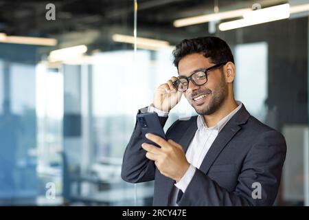Successful financier investor works inside office at work, businessman in business suit uses telephone near window, man smiles and reads good news online from smartphone Stock Photo
