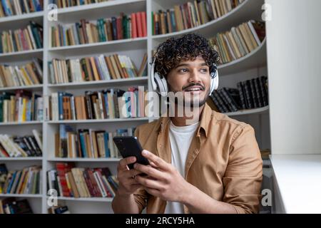 Young hispanic student smiling and happy, using online learning app, smiling man in headphones sitting inside academic library among books. Stock Photo
