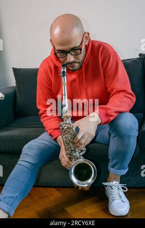 vertical image of male saxophonist with beard, glasses dressed in red, at home thinking about remembering the chords of a song with the saxophone in h Stock Photo