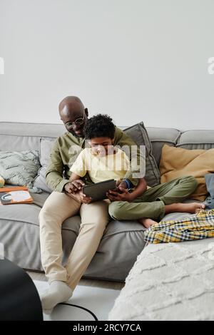 African American dad using digital tablet together with his son while they sitting on sofa in the living room Stock Photo