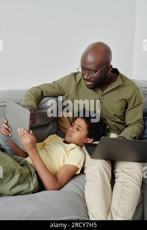 Vertical image of African American dad helping his son to use tablet pc while working on laptop on sofa in the room Stock Photo