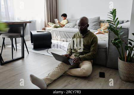 African American father using laptop at his online work at home with his son using tablet pc in background Stock Photo