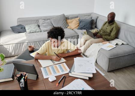 High angle view of African American boy doing homework at table with his dad working on laptop in background Stock Photo