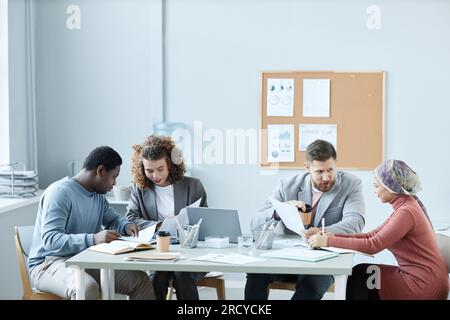 Group of four young intercultural economists discussing financial papers and organizing work while sitting by desk at start-up meeting Stock Photo