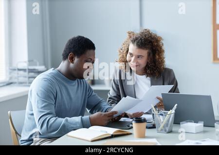 Young African American male broker and his female colleague having discussion of financial data while holding documents at meeting Stock Photo